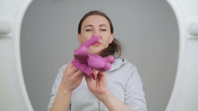 Disgruntled housewife full of determination opens toilet lid, looks through cover inside, puts on pink sanitary gloves , prepares for cleaning and disinfection, . View from toilet bottom. Cleanliness