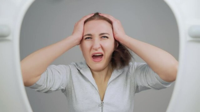 Woman standing in front of urinal holding head in distress or discomfort. Female experiencing moment of discomfort or pain in public restroom