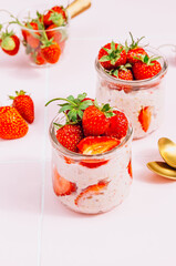 Healthy breakfast of overnight oats with yogurt and fresh strawberries, served in glass jars on a vibrant pink tile table. Bathed in natural light. Selective focus