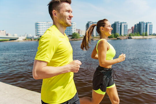 Two people jogging by a river in bright athletic attire, with a modern cityscape in the background.