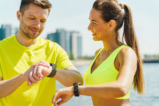 Two joggers in bright sportswear check their fitness watches by the river, with a cityscape behind them. - Powered by Adobe