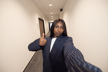 Confident businesswoman in a pinstripe suit taking a selfie in a hallway, giving a thumbs up gesture