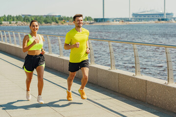 Two joggers in bright athletic wear running along a waterfront path, smiling and enjoying the sunny day.