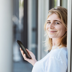 Fototapeta premium A cheerful woman with a smartphone, seen leaning against a wall and smiling. The photo depicts connectivity and happiness.