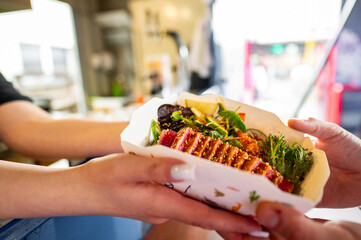 Close-up of hands exchanging a gourmet food with vibrant toppings in a paper tray, suggesting a...