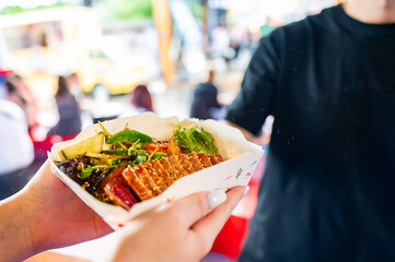 Close-up of hands exchanging a gourmet food with vibrant toppings in a paper tray, suggesting a...