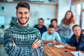 Obraz premium Smiling man in office with colleagues. Smiling young man in a cozy sweater stands confidently in a modern office with his diverse team in the background.