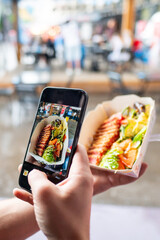 Hands holding a smartphone capture a photo of food with assorted toppings in paper trays. The...