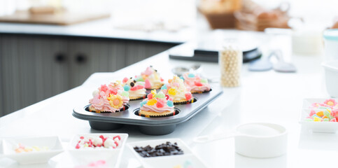 Cupcakes with pastel color butter cream, colorful jelly and chocolate flakes on baking tray in kitchen at home. Flour, sugar, eggs and decorating icing candy on table. Selective focus