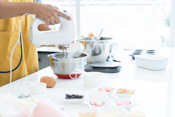 Woman with apron using electric mixer while adding cup of sugar in mixing bowl. Female making cupcakes in kitchen at home. Measuring spoons, scales, sugar, flour, eggs, piping bags are on table