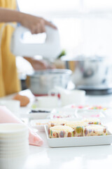 Selective focus on fresh baked cupcakes with ingredients on table. Woman with apron using electric mixer in mixing bowl making cupcakes in kitchen at home at blur background