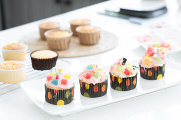 Selective focus on cupcakes with decorations on plate. Fresh baked cupcakes on rack for decorating in paper cups with Icing candy and jelly on white table