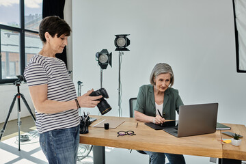 A middle-aged woman sits at a table with laptop, other one with a camera in front of her, ready to capture moments in a professional setting.