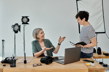Two middle-aged women engage in dialogue while working in front of a laptop in a modern photo...