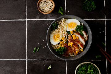 A delicious and hearty bowl of chicken ramen with a boiled egg, fresh green onion, and sweet corn, served over a bed of noodles in a flavorful broth. Black tile background