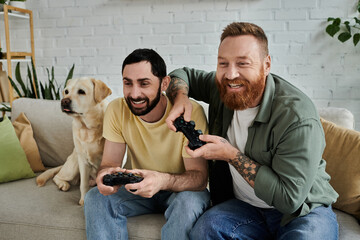 A bearded gay couple and their labrador dog sit on a couch intensively playing a video game together in the living room. © Bliss