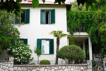 A charming two-story house with green shutters surrounded by lush greenery and well-manicured bushes