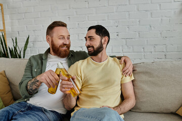 Bearded happy gay couple relaxes on sofa clinking beer bottles at home