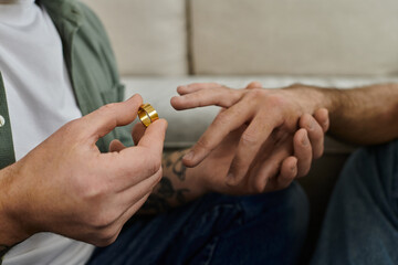 A bearded gay couple sits on a couch, holding hands, man making marriage proposal with ring