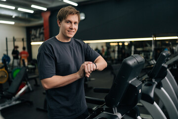 Portrait of cheerful sportsman check health indicator and heart rate on smartwatch during workout strength training, looking at camera. Handsome athlete male using fitness tracker during workout.