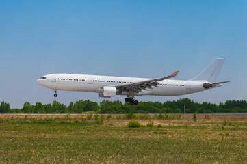 A large white passenger airliner landing on a sunny summer day against the backdrop of a green forest and a cloudless blue sky. Side view.