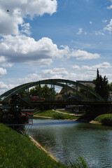 A small iron arch bridge over the river, on which cars and pedestrians drive. The canal is in the city center, people are having lunch at the cafe on the embankment. Town in Serbia is Zrenjanin.