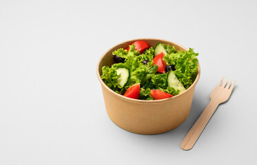 Fresh green salad with tomatoes and cucumbers in a paper container with a wooden fork on a light background. Healthy food concept in biodegradable eco-friendly takeaway dishes. Copy space.