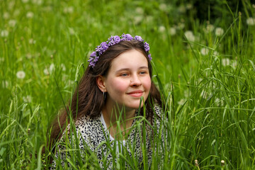 A young girl with flowing hair sits in the green grass in the forest with a purple wreath on her head and smiles.