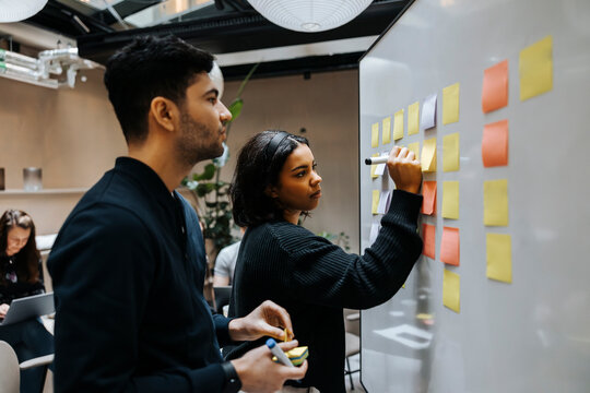 Female entrepreneur writing on adhesive note while standing next to male colleague near whiteboard at office