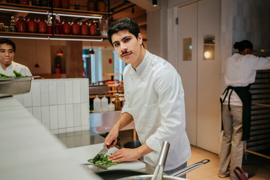 Portrait of male chef cutting broccolini while standing with coworkers in commercial kitchen