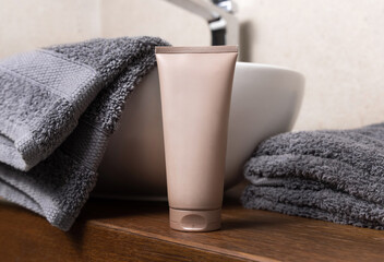Beige cosmetic tube near grey folded towels and basin on wooden countertop in bath, mockup