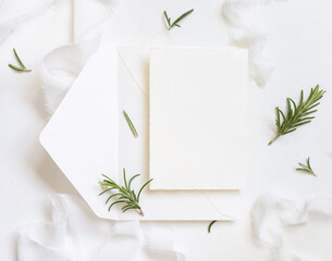 Blank card and envelope near white silk ribbons and rosemary leaves top view, wedding mockup