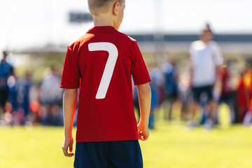 Fototapeta premium Little Boy as a Forward Soccer Player With the Number Seven on His Back. Children Play Football Match Outdoor. School Boy in Red Football Uniform