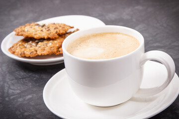Cup of coffee with milk and fresh baked oatmeal cookies with honey and healthy seeds on white plate. Delicious crunchy dessert