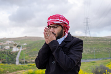 A man in a black jacket and dishdasha stands in a vibrant field of yellow flowers and green hills, arms crossed for warmth, under a cloudy spring sky, feeling cold