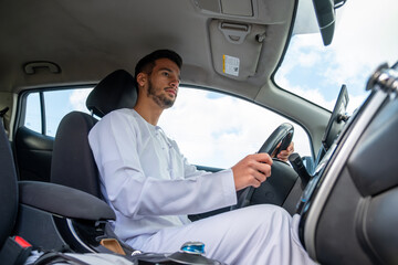 A cheerful young man with a beard, wearing a traditional Middle-Eastern outfit, smiles confidently while seated in his car. He appears to be enjoying a drive through a scenic countryside