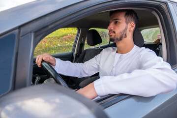 A cheerful young man with a beard, wearing a traditional Middle-Eastern outfit, smiles confidently while seated in his car. He appears to be enjoying a drive through a scenic countryside