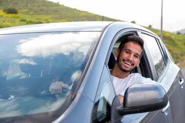 A cheerful young man with a beard, wearing a traditional Middle-Eastern outfit, smiles confidently while seated in his car. He appears to be enjoying a drive through a scenic countryside