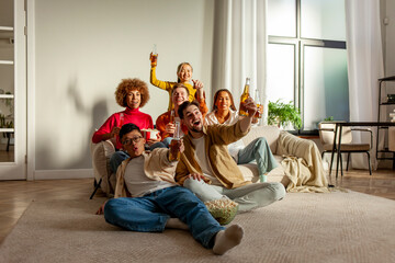multiracial group of young friends sitting on the couch at home drinking beer and watching TV and celebrating good luck