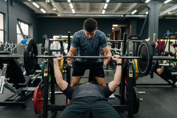 Portrait of personal fitness trainer helping beginner sportsman doing barbell bench press exercise...