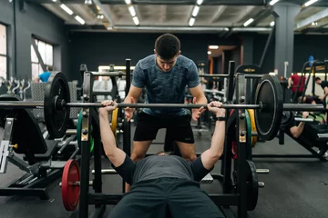 Fotobehang Persoonlijk Portrait of personal fitness trainer helping beginner sportsman doing barbell bench press exercise during workout in gym. Athletic male doing barbell bench press under couch supervision.  © dikushin