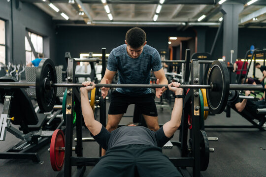 Portrait of personal fitness trainer helping beginner sportsman doing barbell bench press exercise during workout in gym. Athletic male doing barbell bench press under couch supervision.
