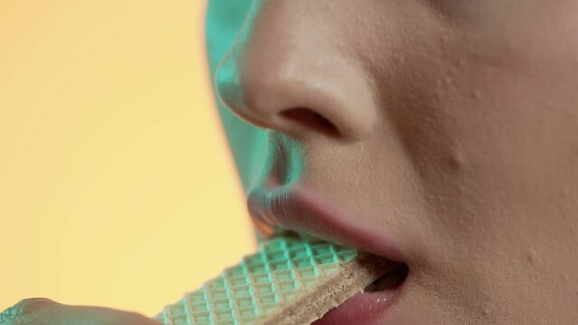 Makro zbliżenie młodej kobiety bez makijażu jedzącej wafelka i oblizującej usta na ż&oacute;łtym tle z zielonym światłem &mdash; slow motion 4K, naturalność, smak i kobiecość