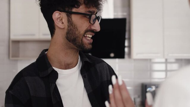 Young curly man in glasses shouting at a young woman and gesticulating in the kitchen at home during a quarrel close up. Concept of problems in the relationship of a couple