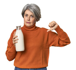 Caucasian mid-age female holding milk bottle showing a dislike gesture, thumbs down. Disagreement concept.