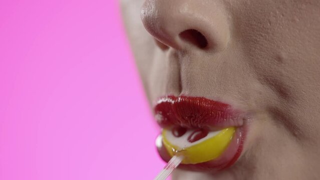Makro ujęcie kobiety na r&oacute;żowym tle. Ma czerwoną szminkę, bierze do ust kolorowy lizak z wzorem czereśni, oblizuje go zmysłowo, wyjmuje i namiętnie oblizuje usta, kończąc uśmiechem