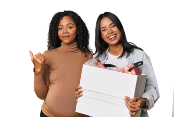 Latin women couple moving, one pregnant  smiling and pointing aside, showing something at blank space.