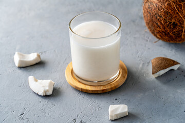 A refreshing glass of coconut milk surrounded by coconut pieces on a textured gray surface,...