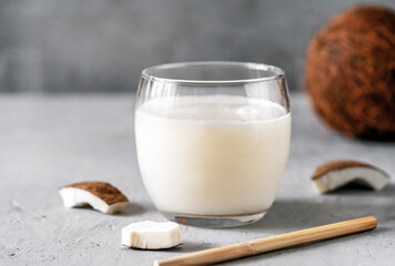 A glass of coconut milk surrounded by coconut pieces on a textured gray background, highlighting a natural and healthy beverage.