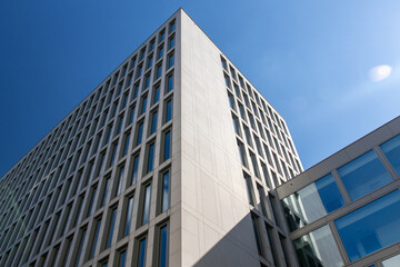 Fototapeta premium A photo of the corner of a modern, multi-story building with a lattice-like window design, framed against a clear blue sky. The building is constructed of light-colored materials.